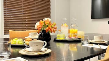 Una mesa de desayuno servida con croissants, fruta y bebidas, en una habitación moderna con televisor y persianas.