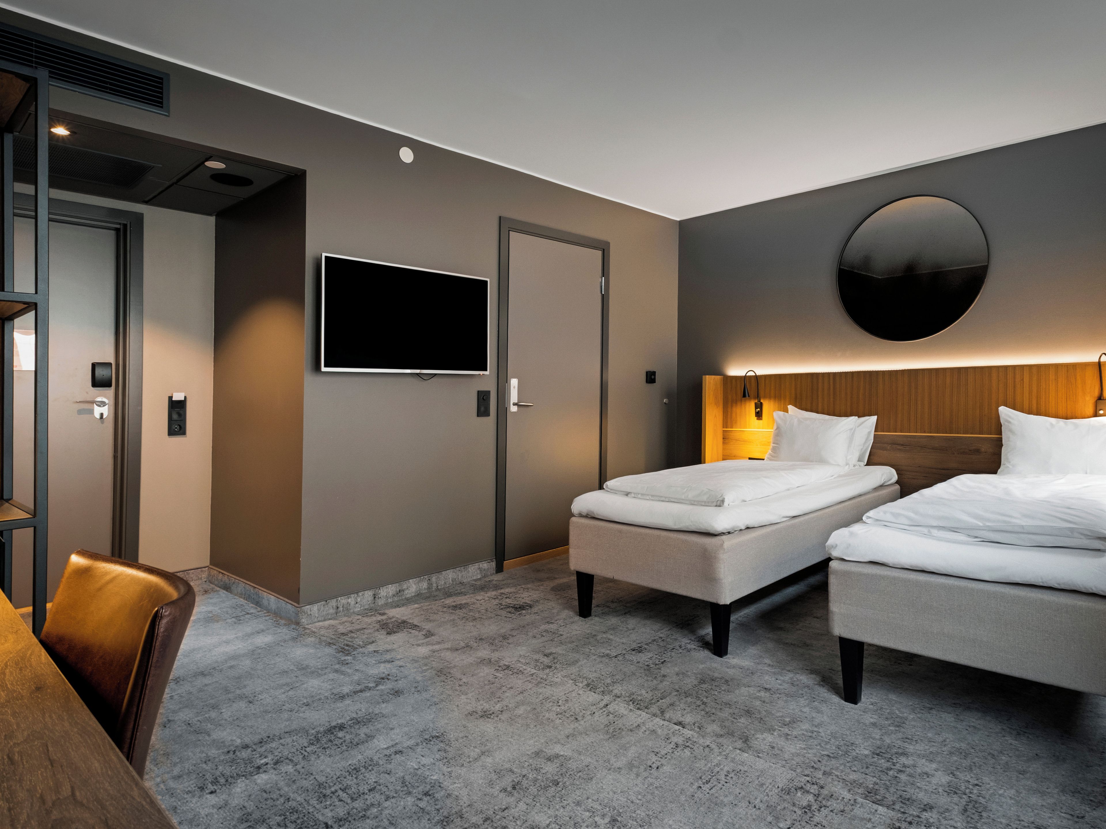 A hotel room with two beds, a flat-screen TV, a round wall-mounted light, and a wooden headboard.