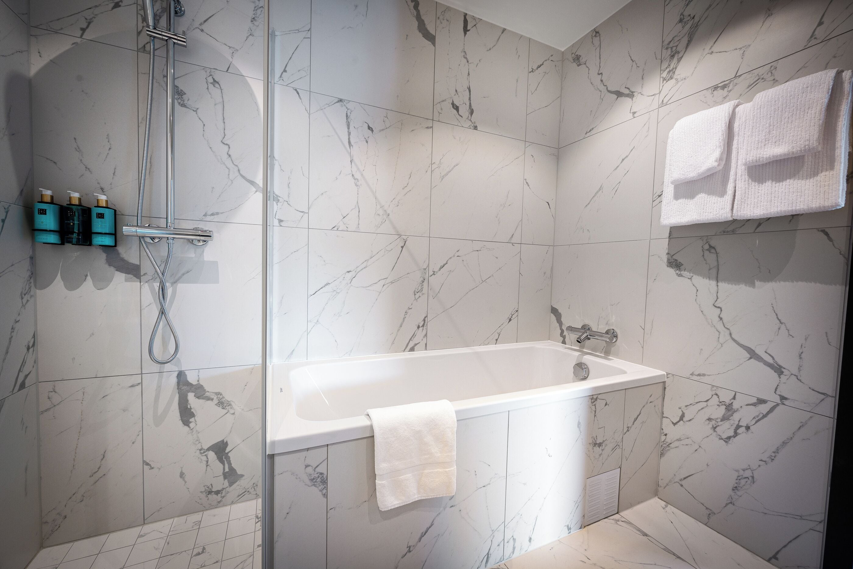 A modern bathroom with a marble shower wall, a white bathtub, and a towel rack with two towels.
