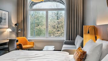 A hotel room with a large bed, a desk, a chair, and a view of trees through a window.