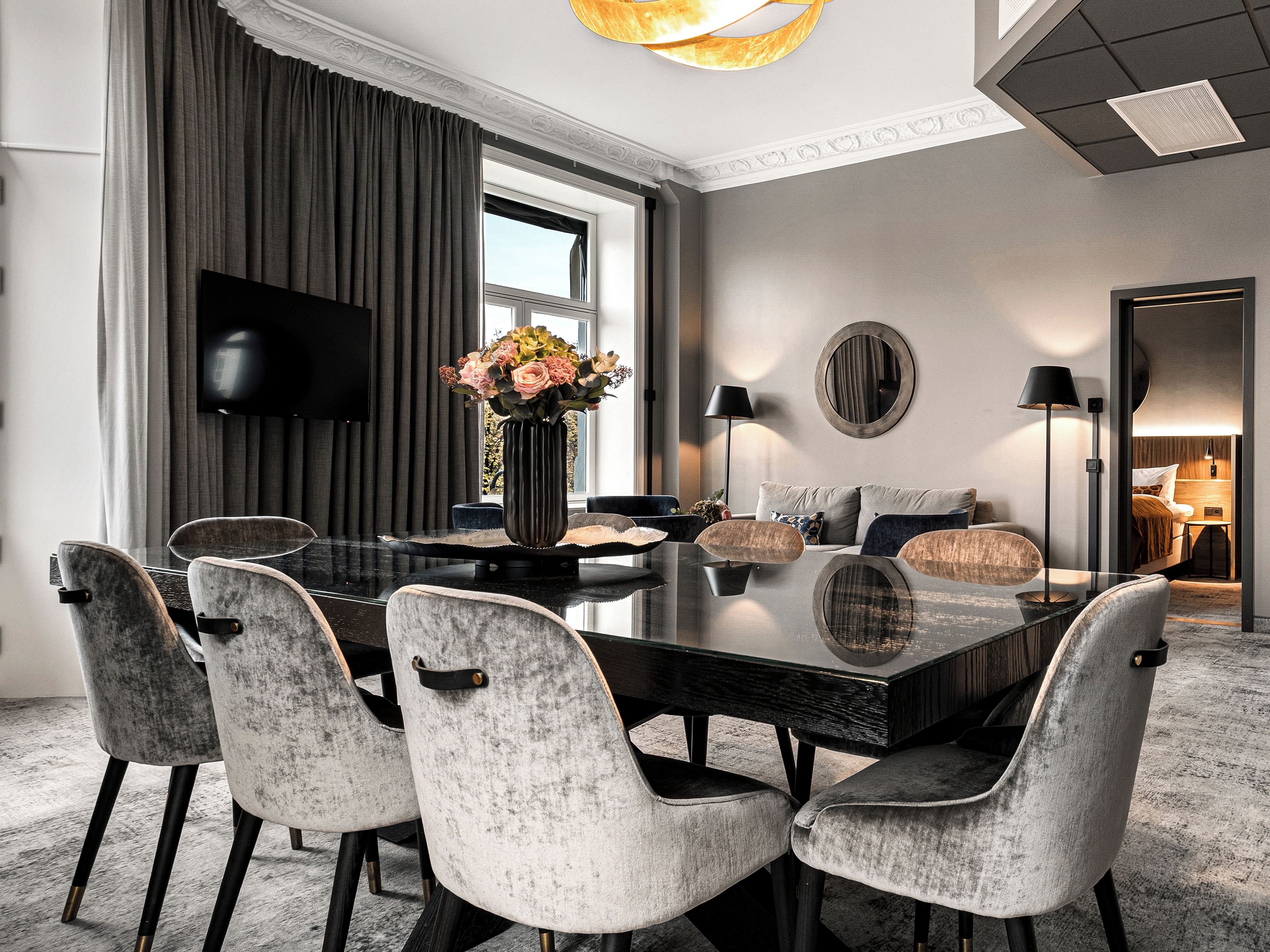 Un espace repas moderne avec une table en verre, des chaises à revêtement gris, un vase avec des fleurs et un téléviseur à écran plat fixé au mur.