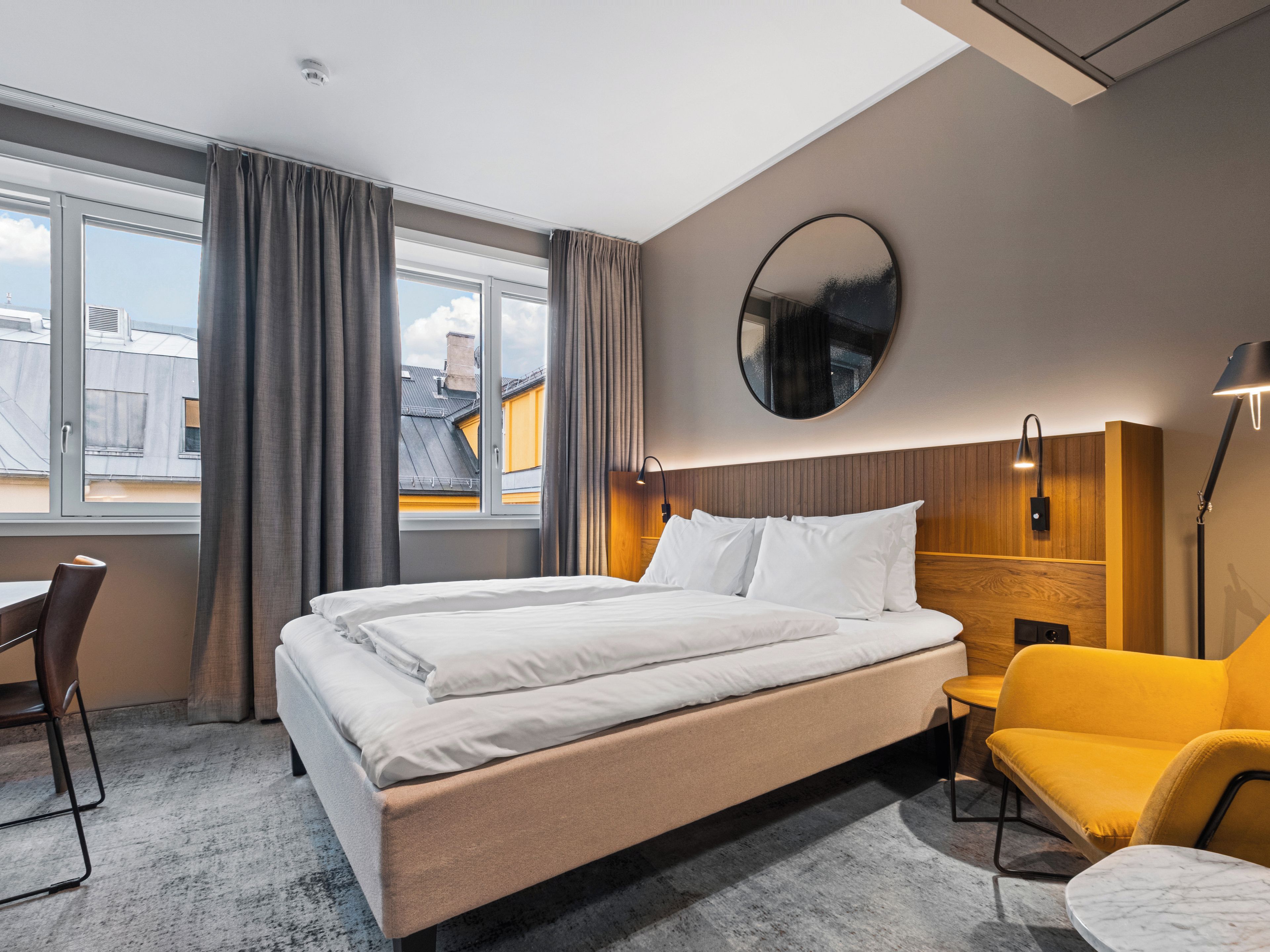 A hotel room with a large bed, a round mirror, a yellow armchair, a small table, and a view of buildings through the window.