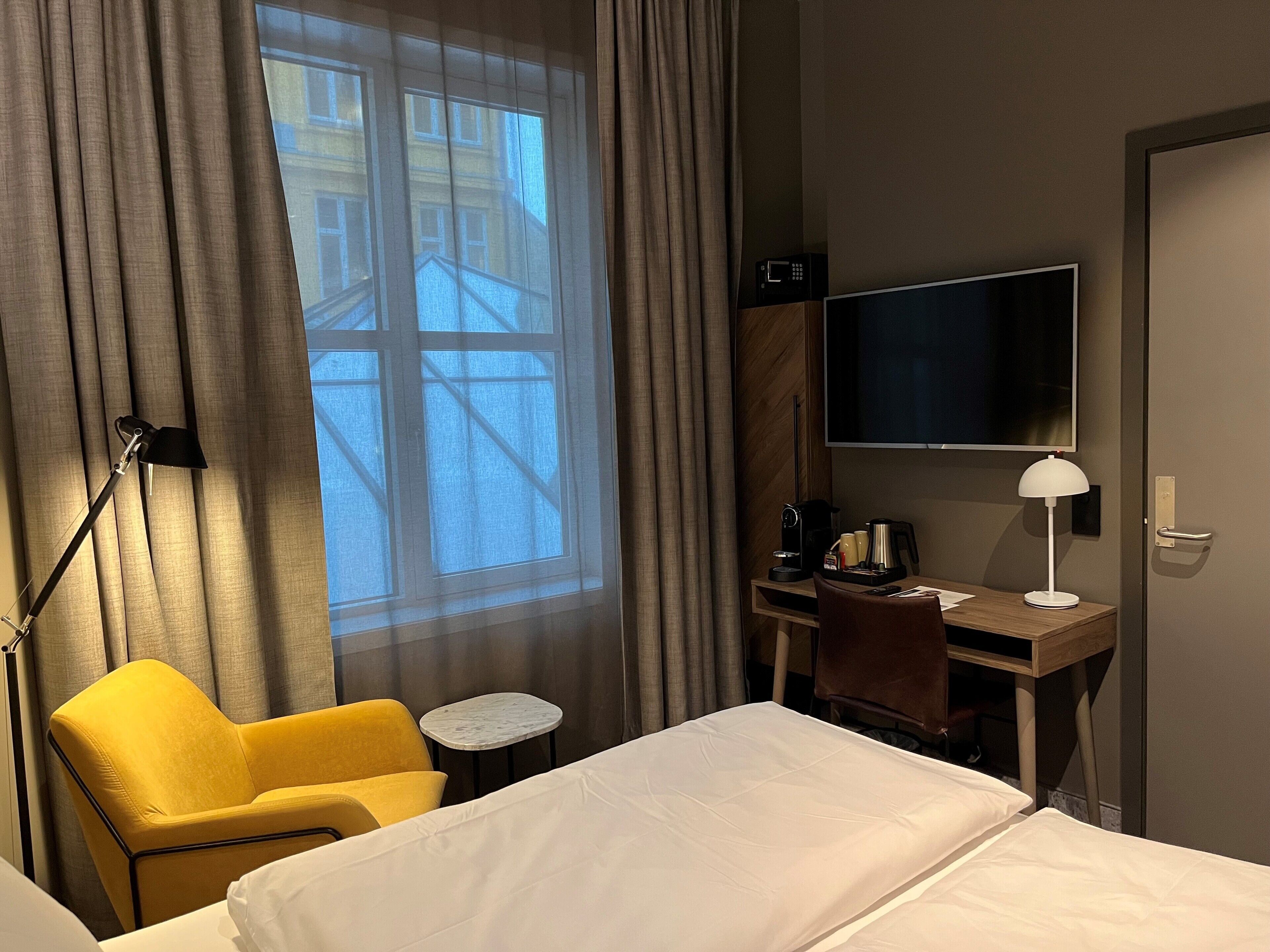 Habitación de hotel con cama, escritorio con computadora, silla, mesita pequeña, lámpara y televisor.