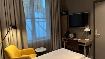 Habitación de hotel con cama, escritorio con computadora, silla, mesita pequeña, lámpara y televisor.
