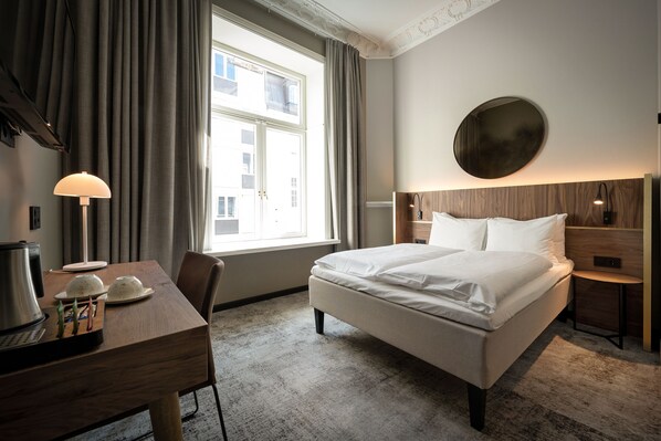 A hotel room with a large bed, a desk with a lamp, a coffee maker, and a view of the city through the window.