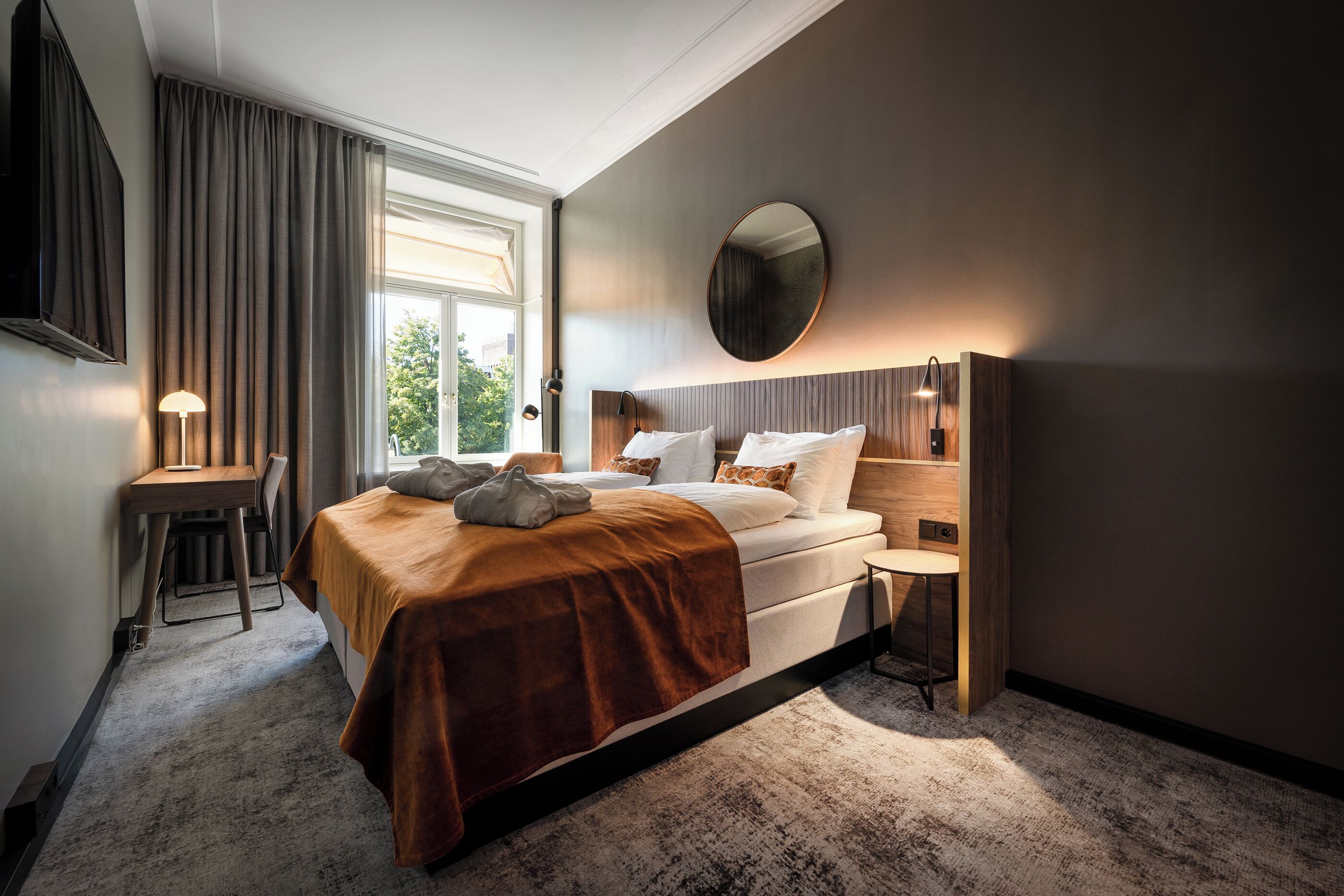 Quarto de hotel moderno com uma cama grande, uma escrivaninha e um espelho redondo.
