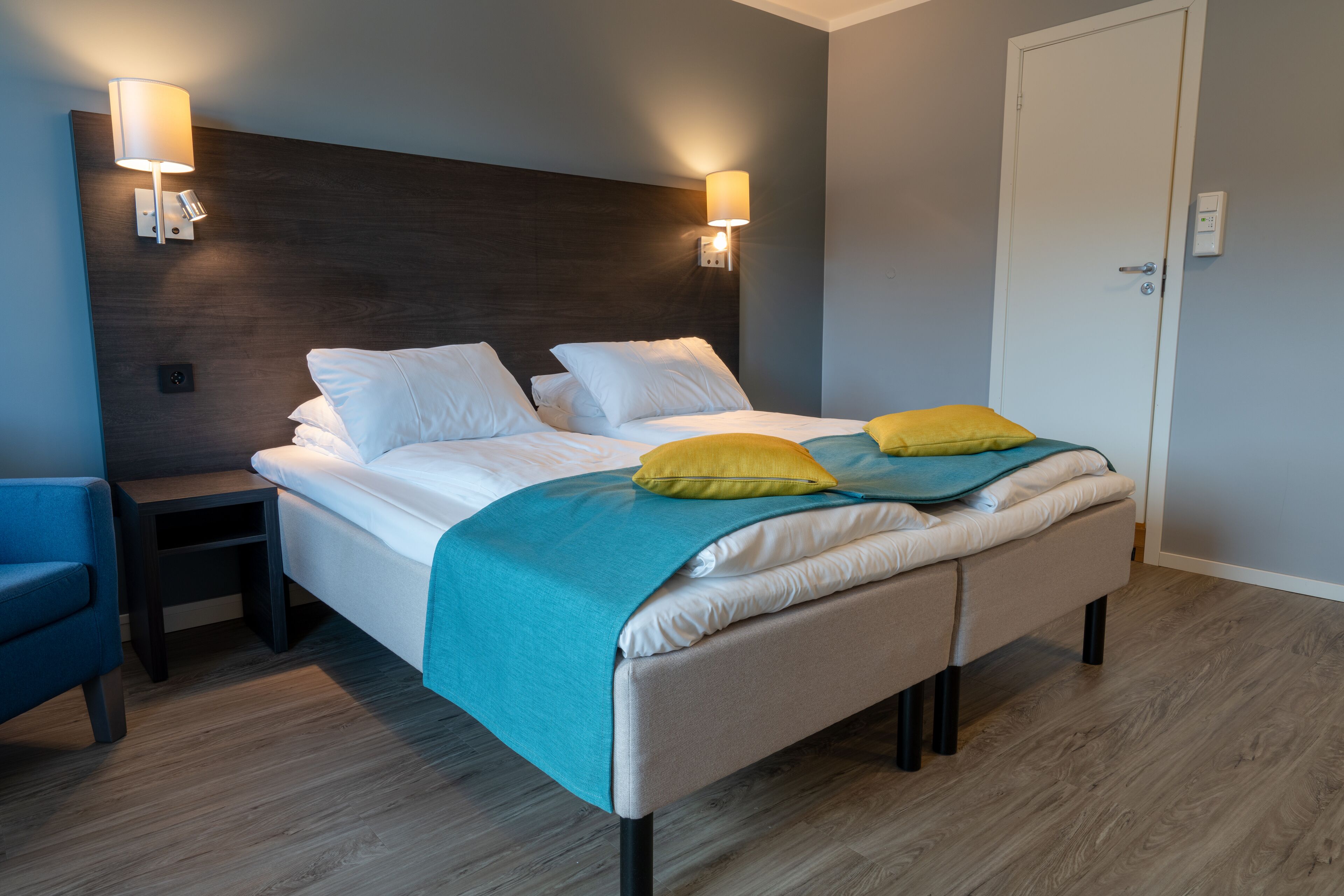 A modern hotel room with a large bed, two bedside lamps, a blue armchair, and a wooden floor.