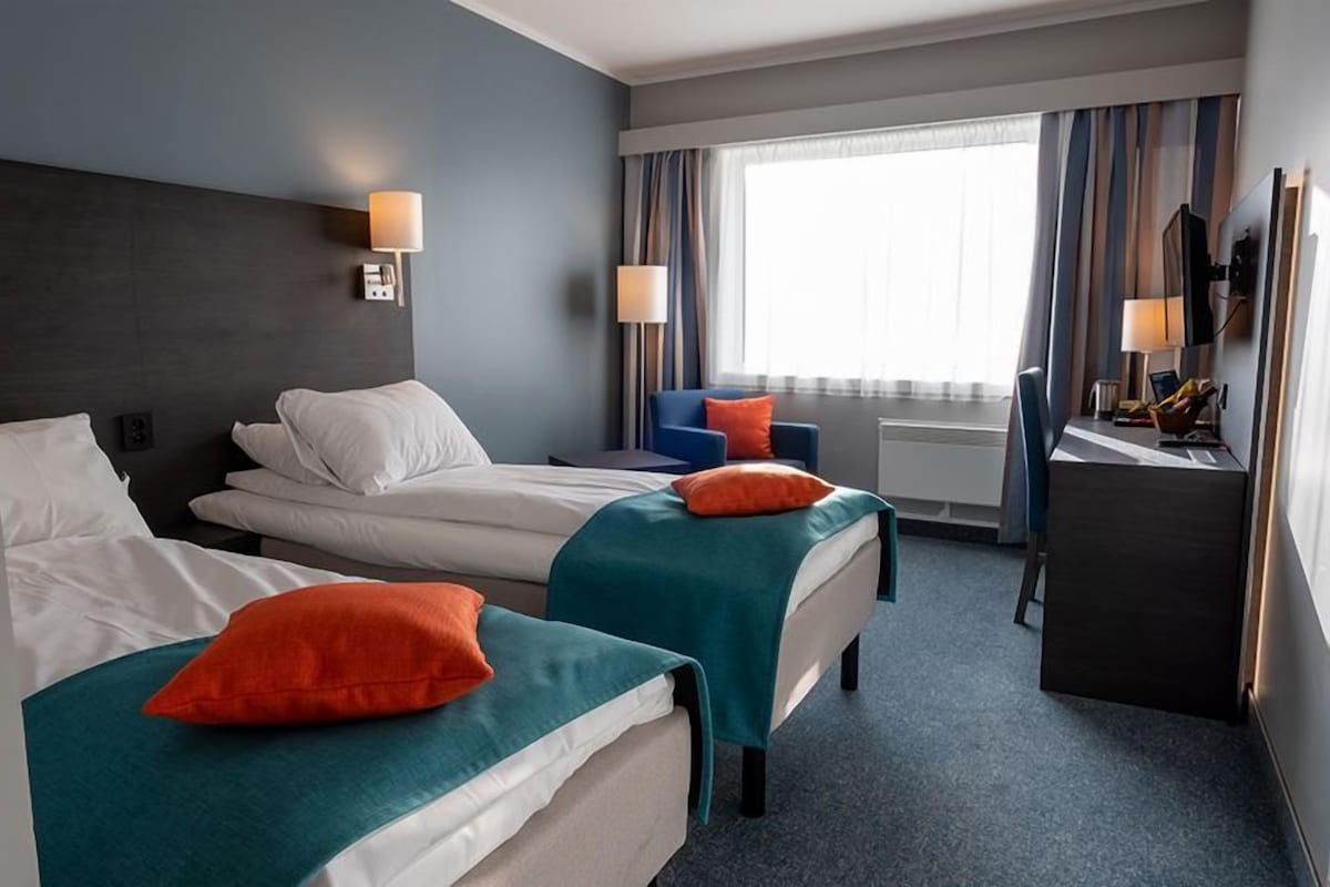Una habitación de hotel con dos camas, un escritorio, una silla y un ventanal con cortinas.