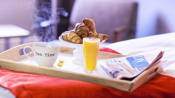 Un plateau de petit-déjeuner avec des croissants, une tasse portant l’inscription « Heure du thé » et un verre de jus d’orange, posé sur un lit recouvert d’une couverture rouge.