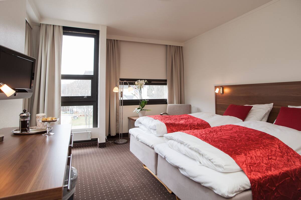 A hotel room with two beds, a TV, a desk with a coffee maker, and a window with curtains.