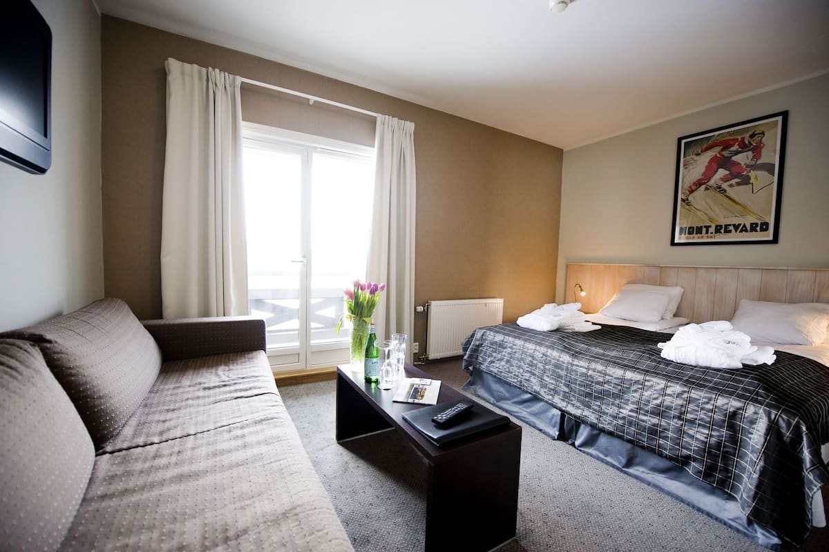 A hotel room with a bed, a sofa, a small table with a vase of flowers, a radiator, and a framed picture on the wall.