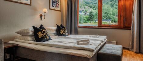 Habitación de hotel con una cama grande, una mesita de noche, una lámpara, dos obras de arte enmarcadas, una ventana con vistas a un bosque y un banco.