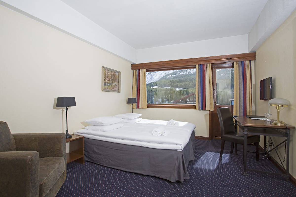 A hotel room with a bed, a desk, a chair, a lamp, a TV, and a window with curtains.