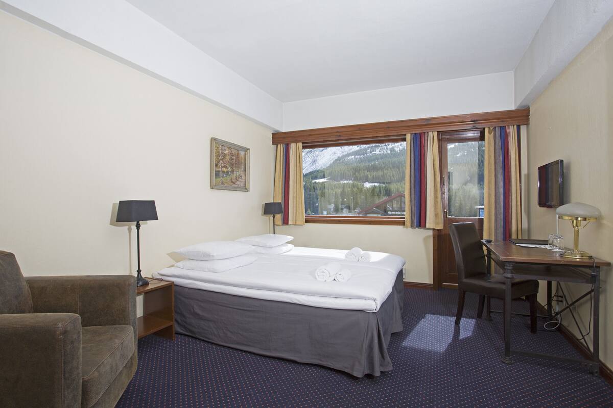 A hotel room with a bed, a desk, a chair, a lamp, a TV, and a window with curtains.