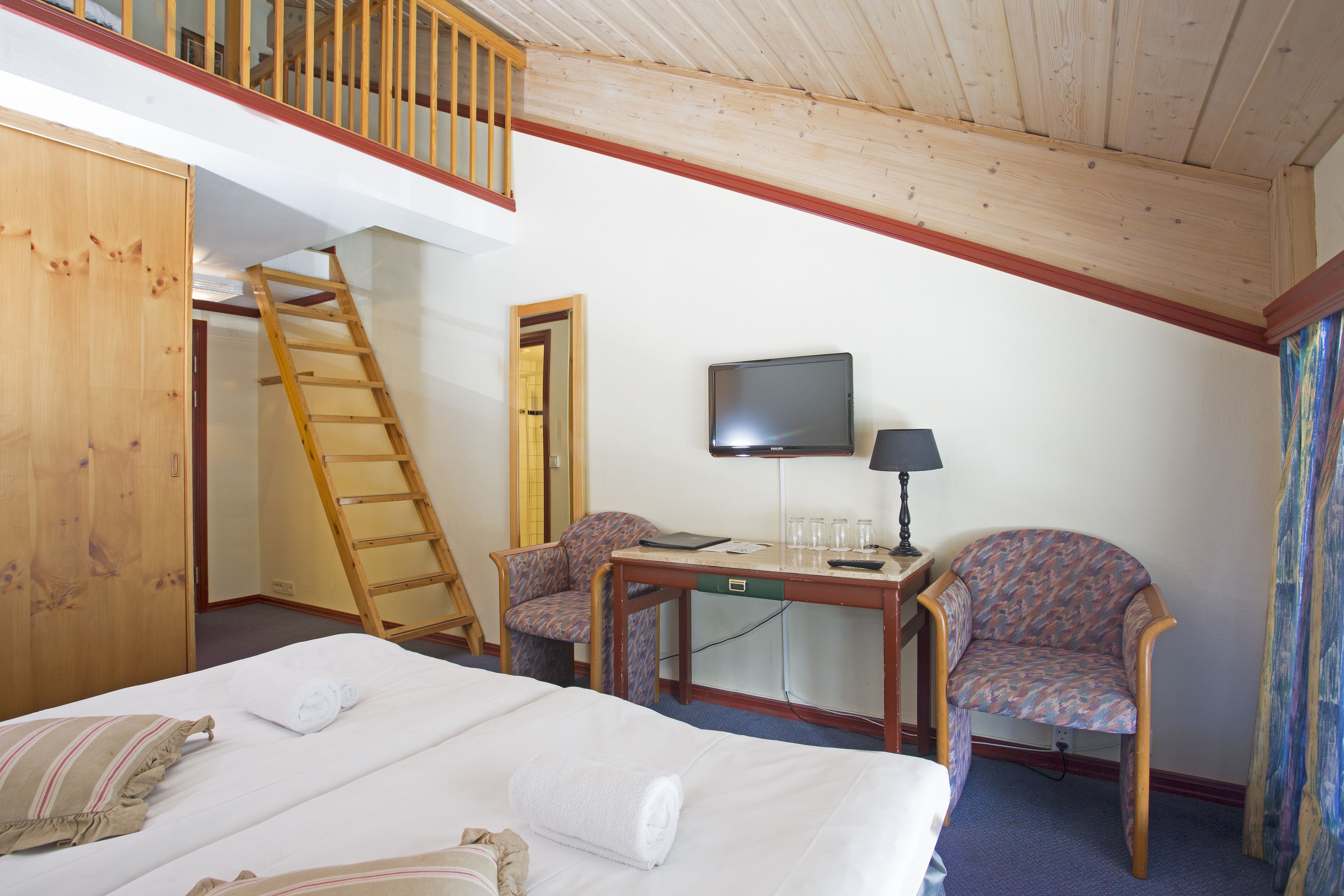 A hotel room with a bed, two chairs, a desk, a television, and a wooden staircase.
