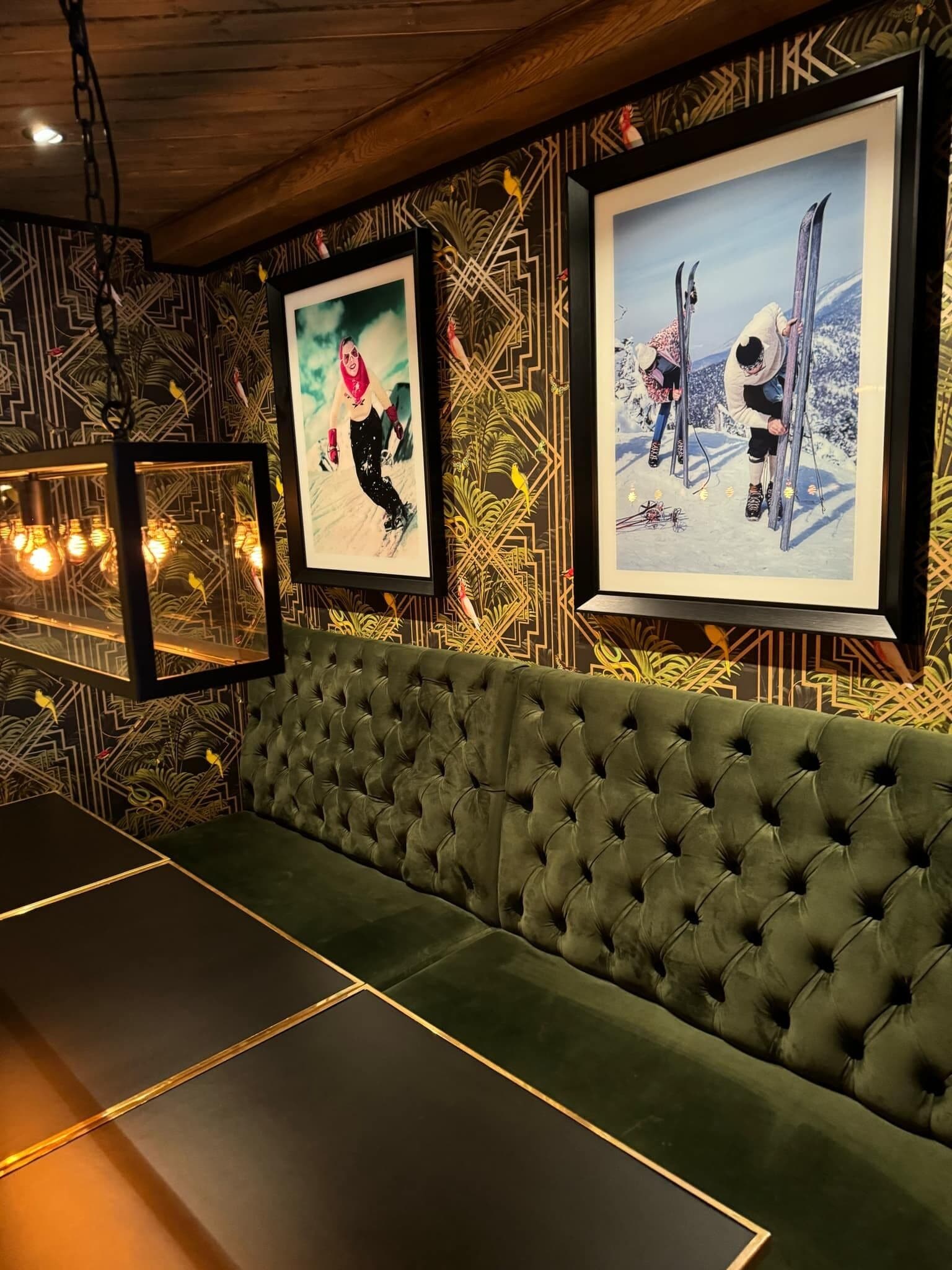 Een groene sofa met losse kussens, tegen een muur met behang met een geometrisch patroon en ingelijste kunstwerken.