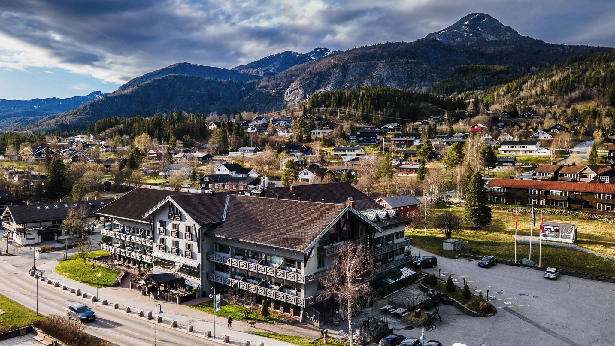 Et hotel med en stor bygning, parkeringsplads og et bjerglandskab i baggrunden.