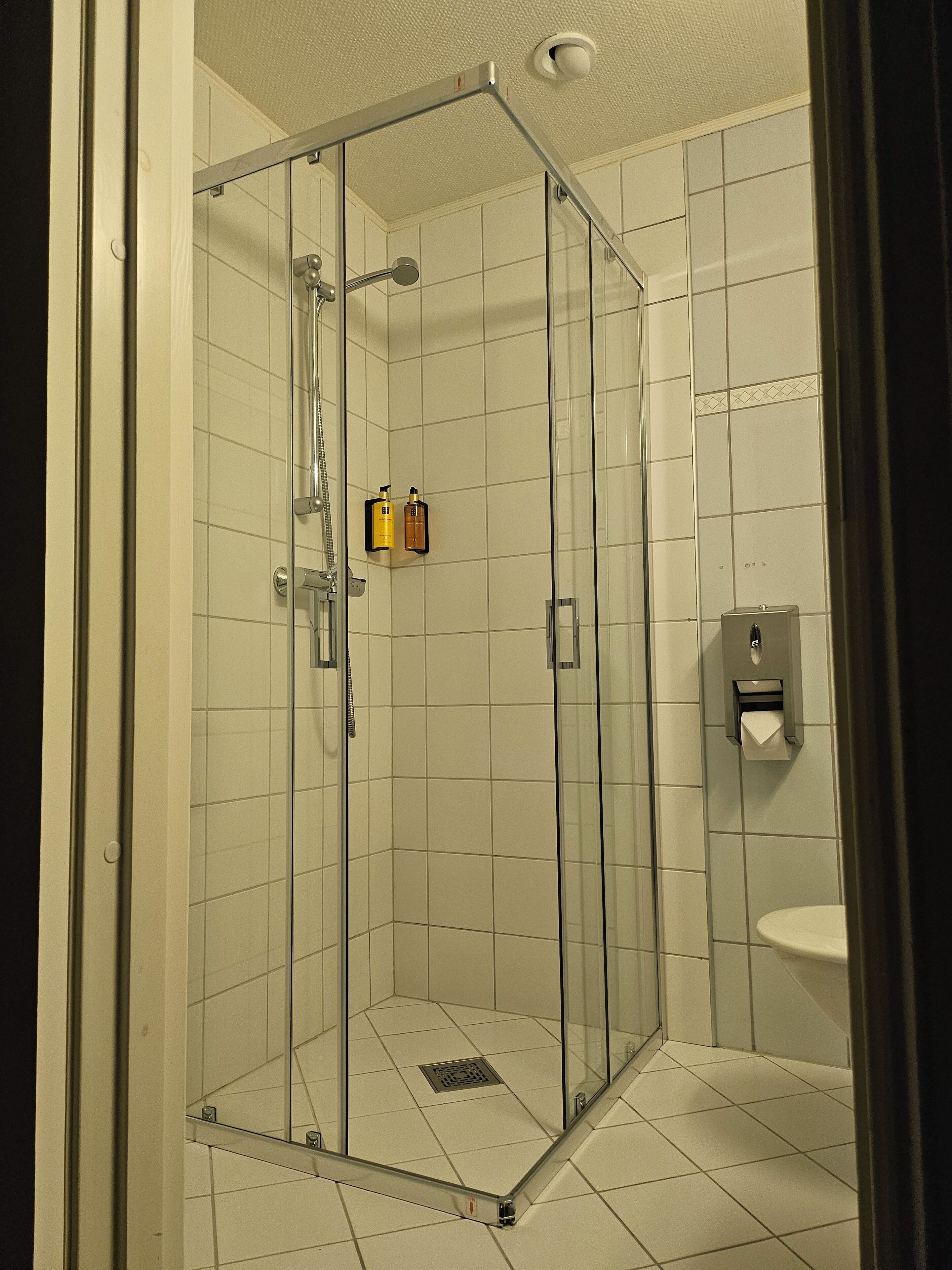 A glass-enclosed shower stall with a white tiled floor and a wall-mounted soap dispenser.