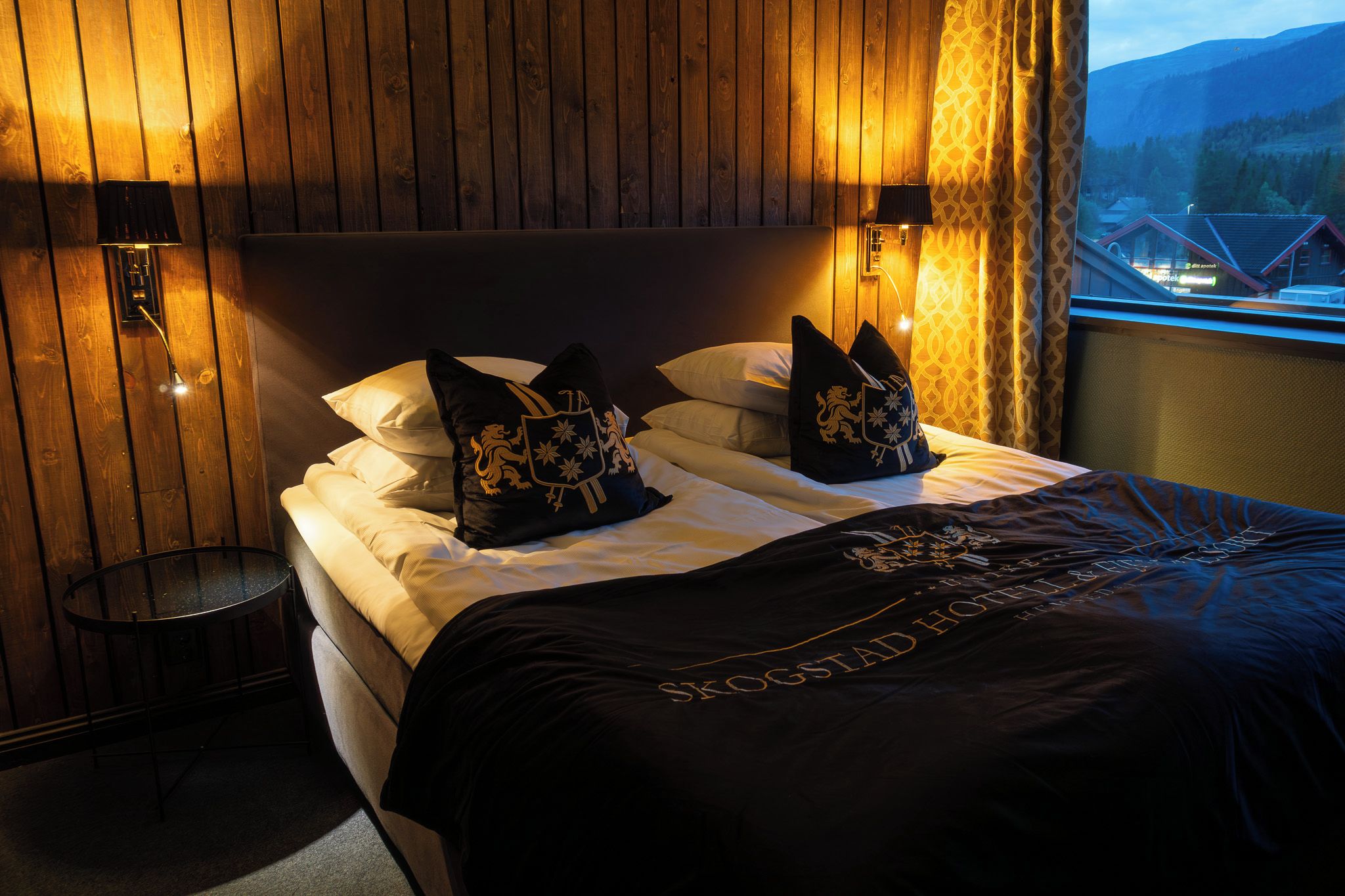 A hotel room with a wooden headboard, two bedside lamps, a bed with white linens and black pillows with gold emblems, and a view of a mountain landscape through a window.