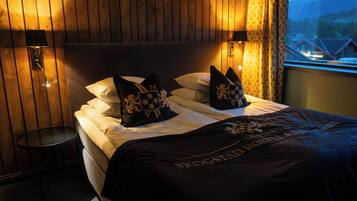 A hotel room with a wooden headboard, two bedside lamps, a bed with white linens and black pillows with gold emblems, and a view of a mountain landscape through a window.