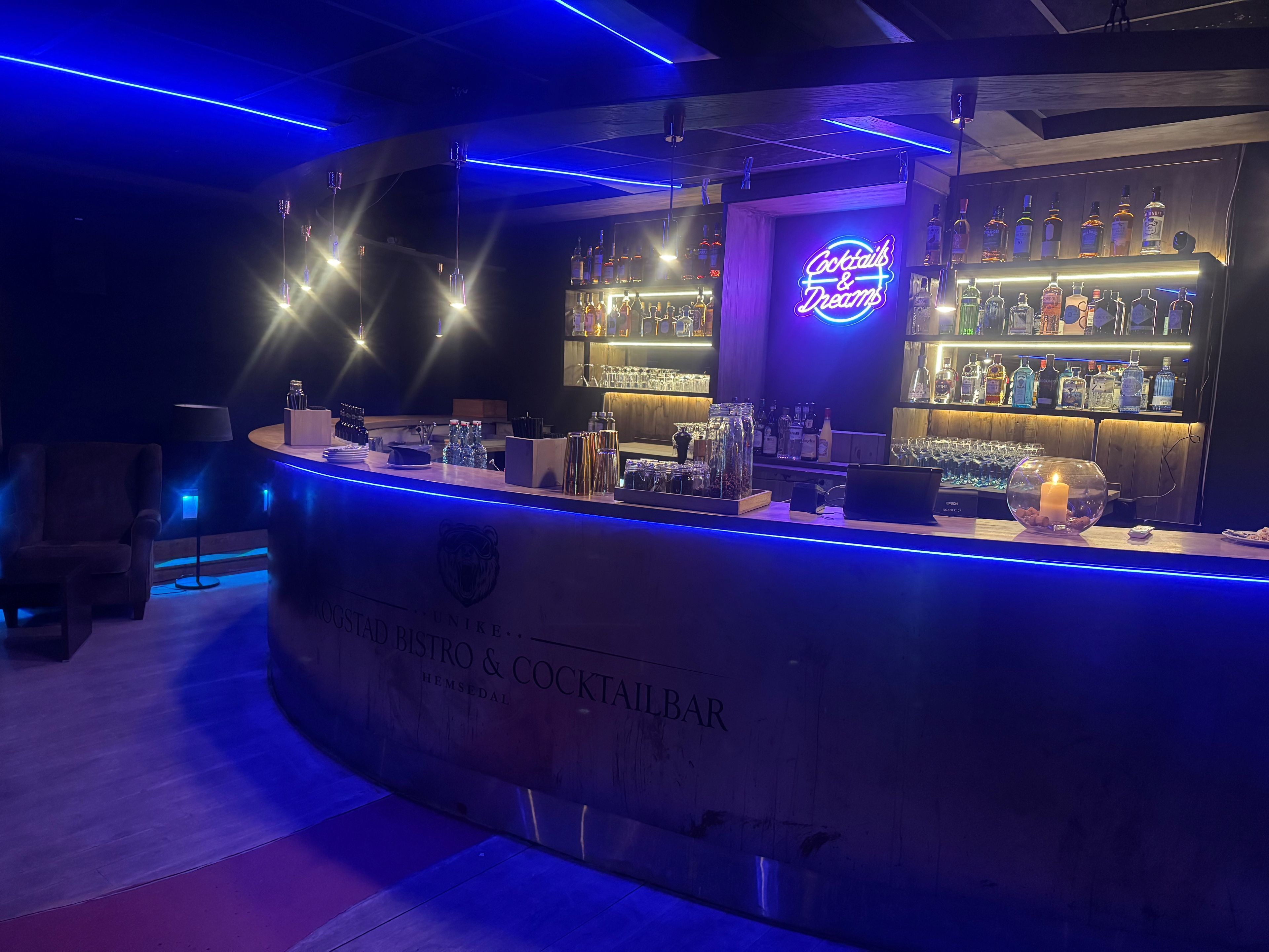 A bar area with a neon sign, shelves of liquor bottles, and a counter with various bar tools.