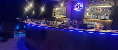 A bar area with a neon sign, shelves of liquor bottles, and a counter with various bar tools.