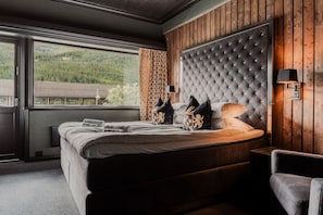 A bedroom with a large bed, a wooden headboard, a nightstand, a lamp, and a view of a forest through a window. - Skogstad Hotell - Unike Hoteller (Hemsedal)