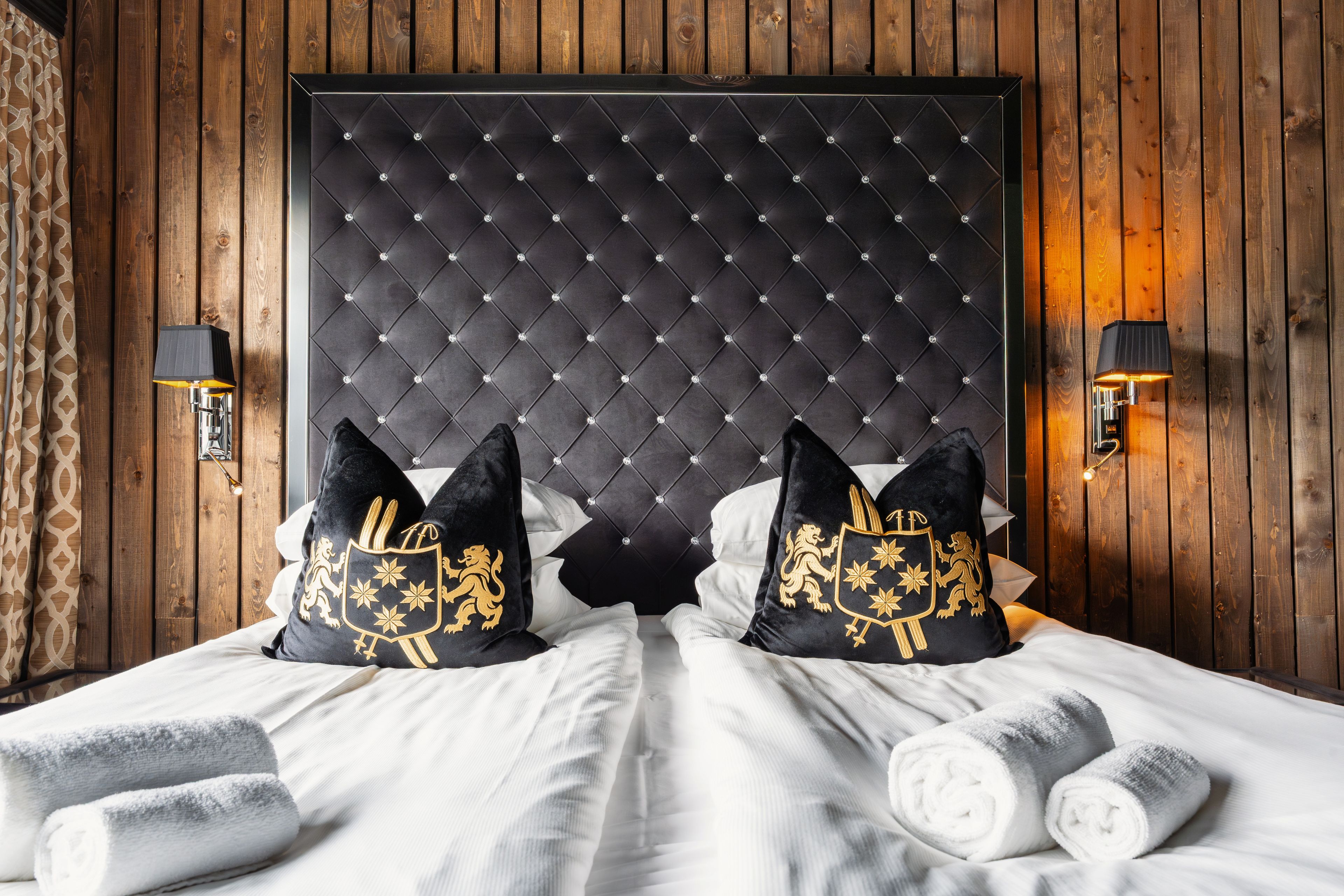 A hotel room with a tufted headboard, two decorative pillows with gold emblems, a bed with white linens, and two wall-mounted lamps.