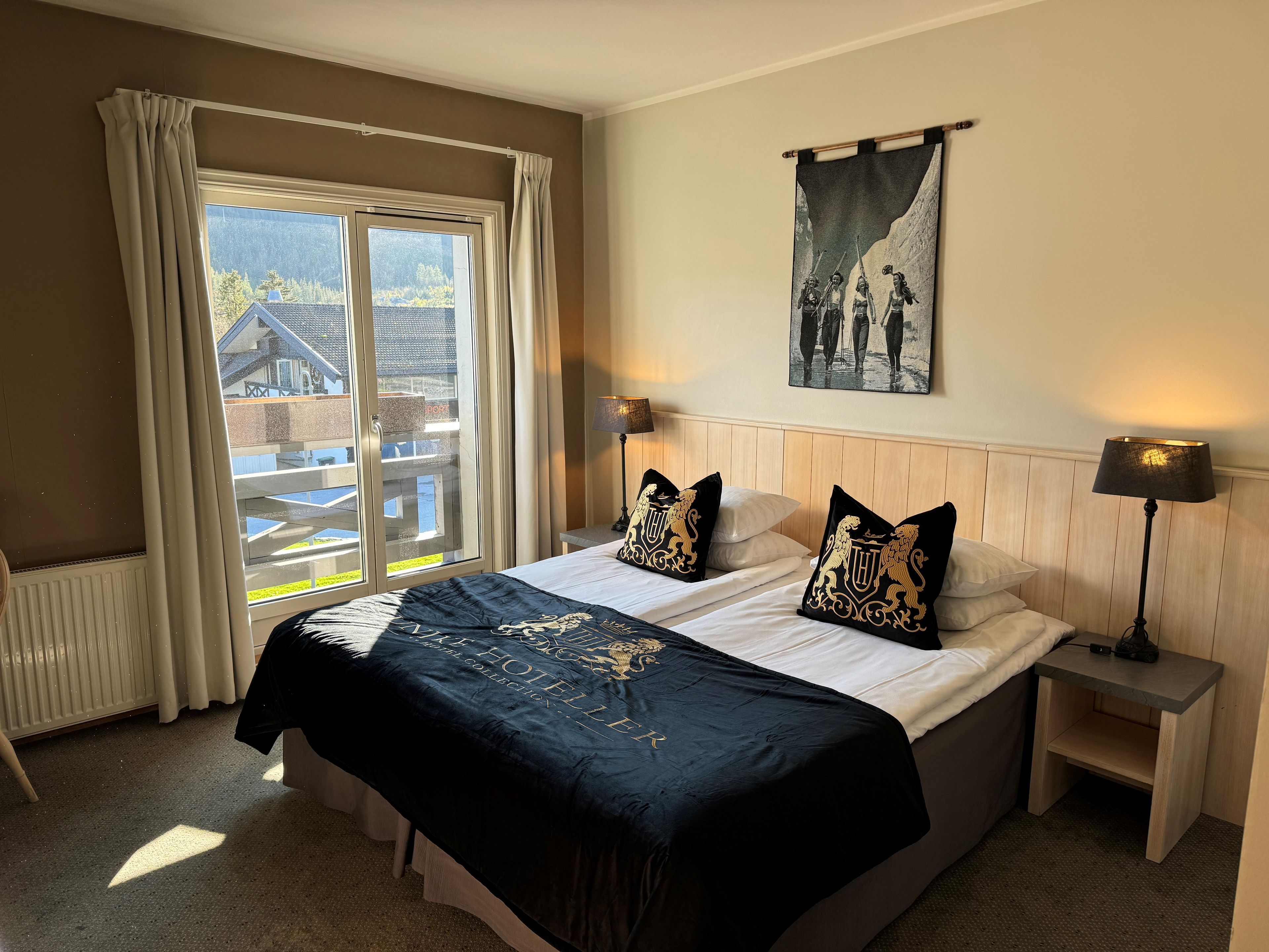A hotel room with a bed, two bedside tables, a window with curtains, a wall-mounted picture, and a radiator.