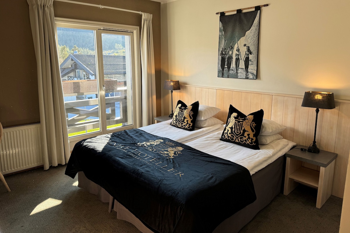 Habitación de hotel con cama, dos mesitas de noche, ventana con cortinas, un cuadro colgado en la pared y un radiador.