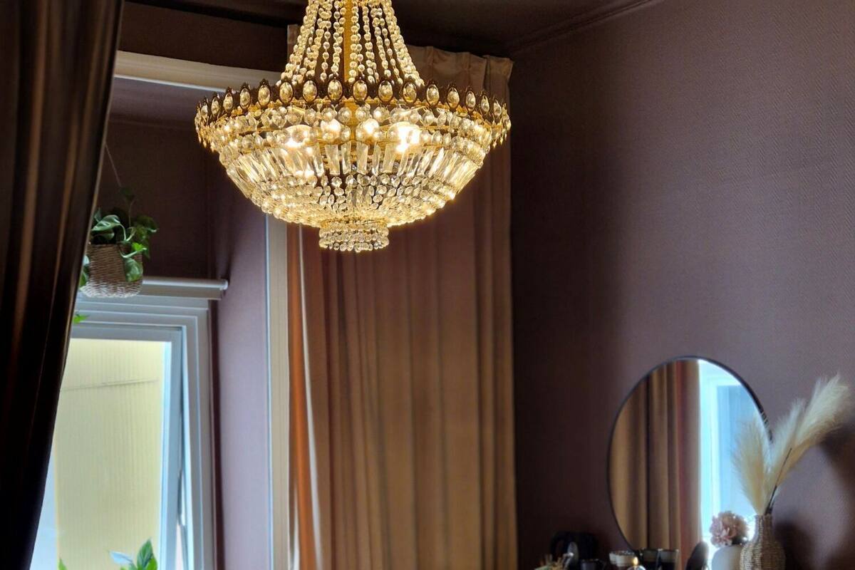 Quarto com cama grande, lustre, penteadeira, espelho e janela com cortinas.