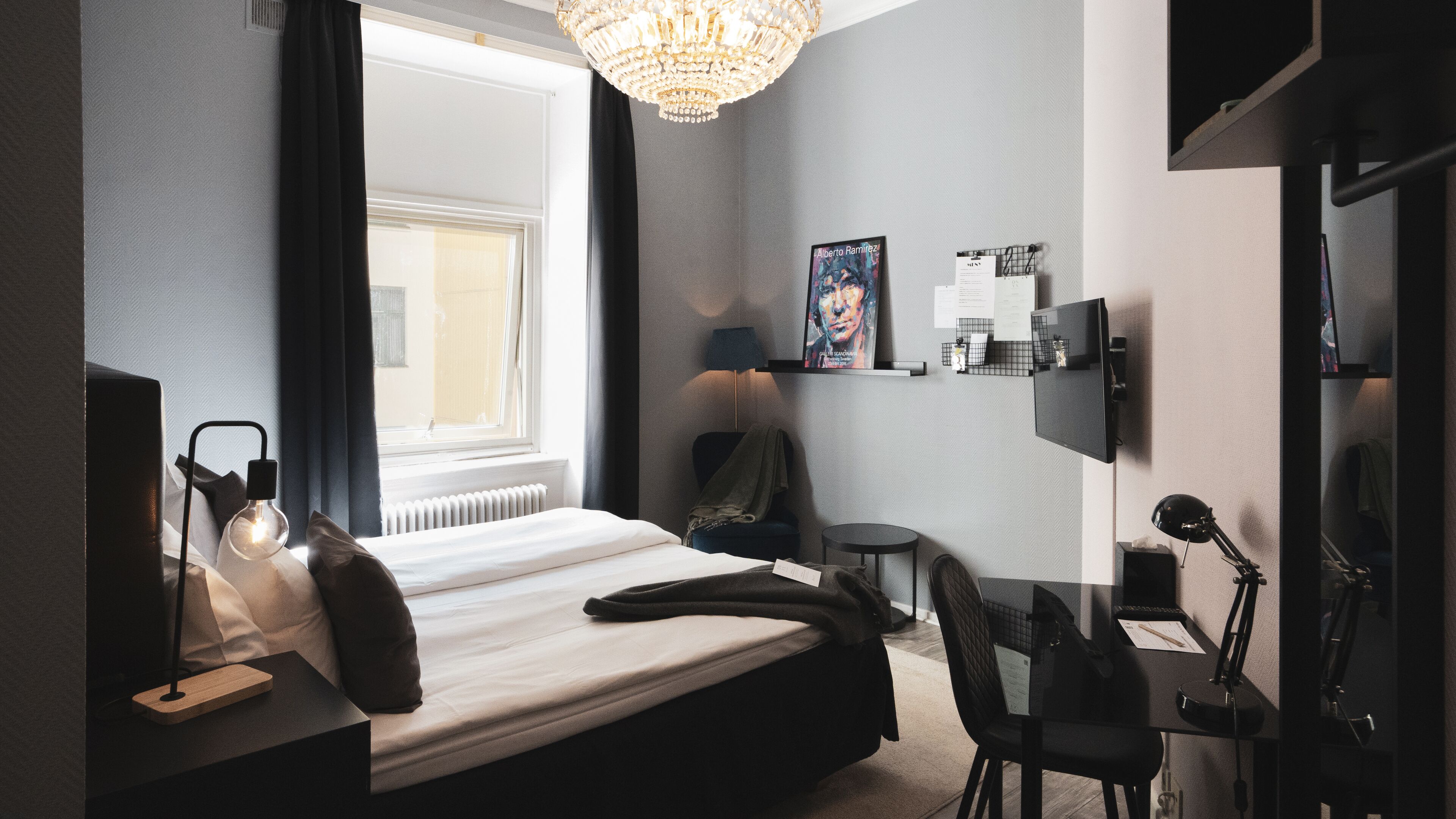Hotellihuone, jossa on sänky, työpöytä, tuoli, televisio, lamppu ja ikkuna, jossa on verhot.