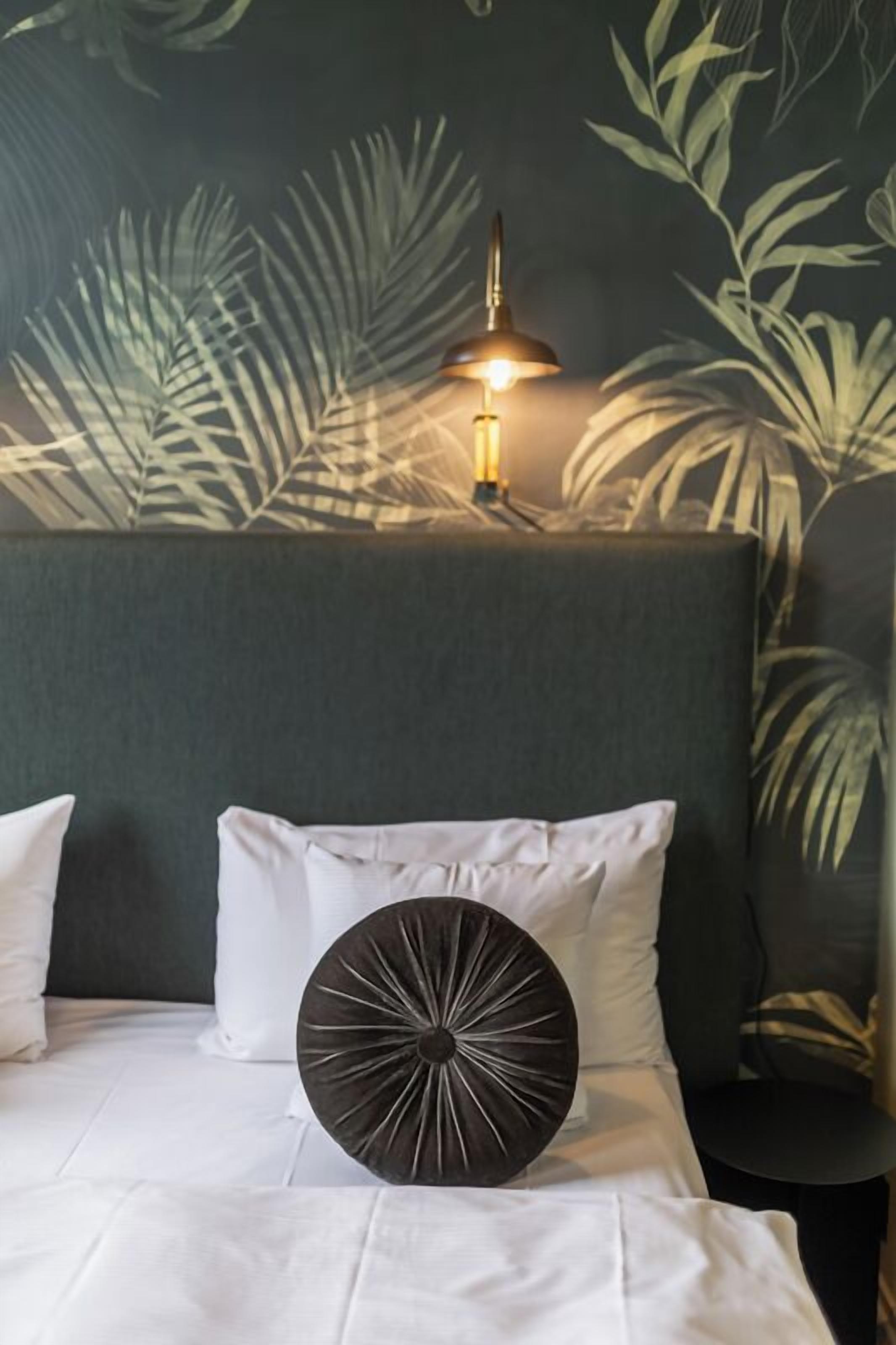 Una cama con ropa blanca, un cojín decorativo redondo oscuro y una lámpara de pared con luz encendida.