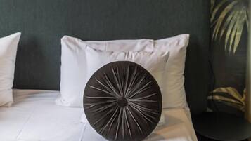 Una cama con ropa blanca, un cojín decorativo redondo oscuro y una lámpara de pared con luz encendida.