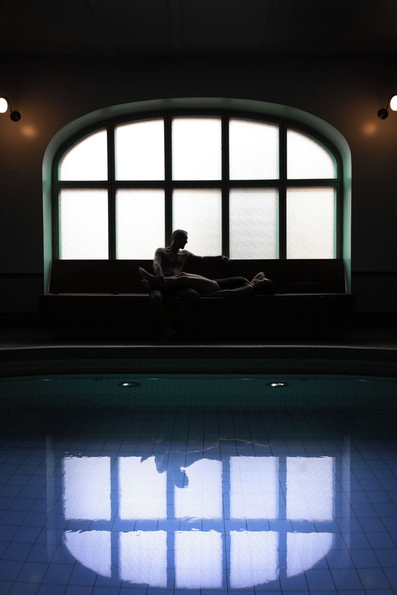 A person sitting on a bench by a large arched window with a reflection in the pool below.