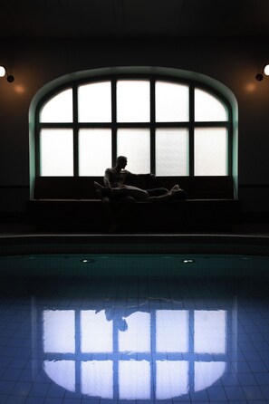 Una persona sentada en un banco junto a un ventanal arqueado, con su reflejo en la piscina de abajo.