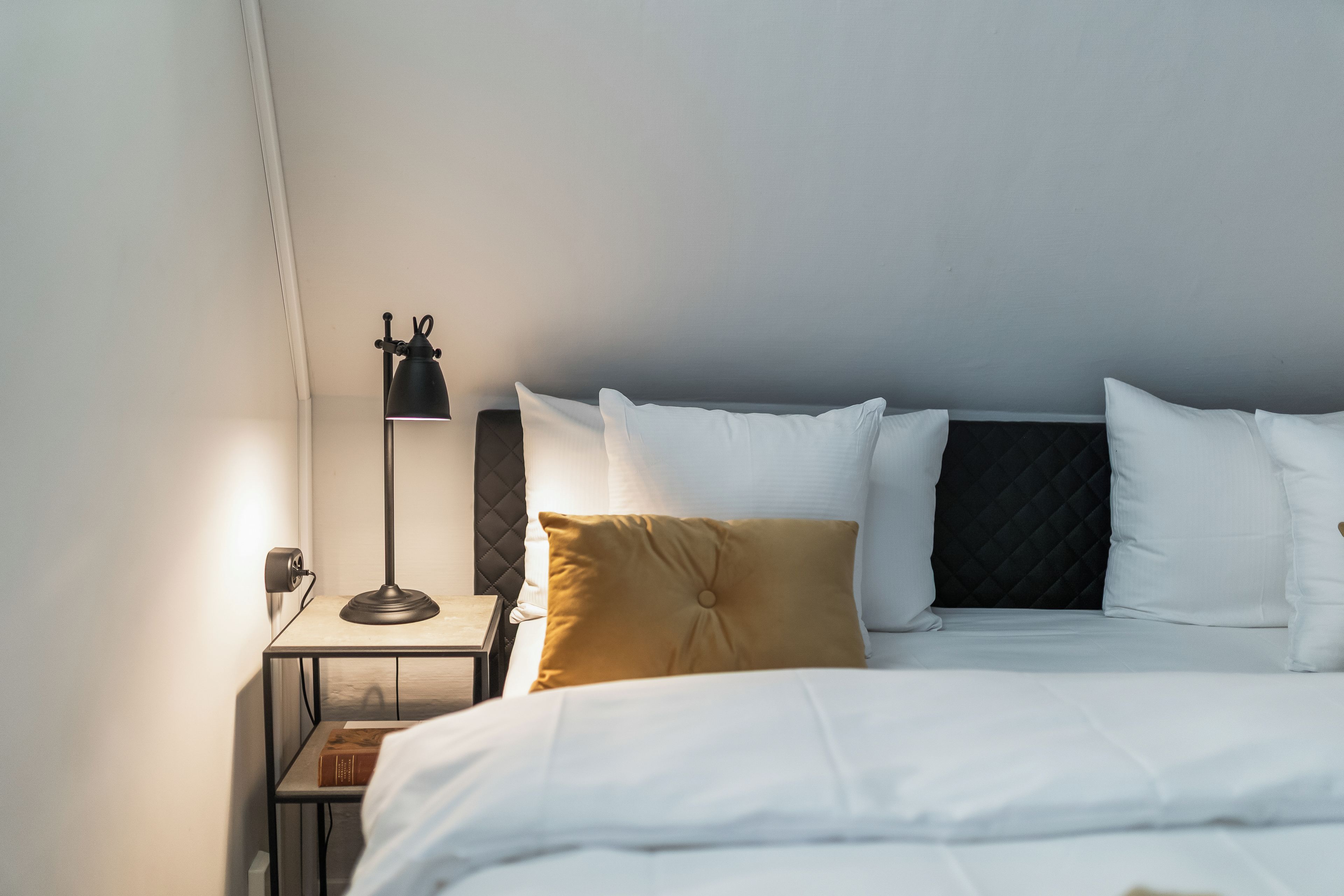 Una cama bien hecha con sábanas blancas, una sola almohada marrón y un cabecero negro. Una lámpara de pie con pantalla negra está ubicada a la izquierda de la cama.