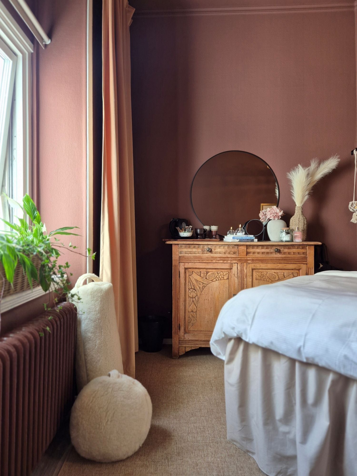 A bedroom with a bed, a dresser, a mirror, a radiator, a window with curtains, and a potted plant.