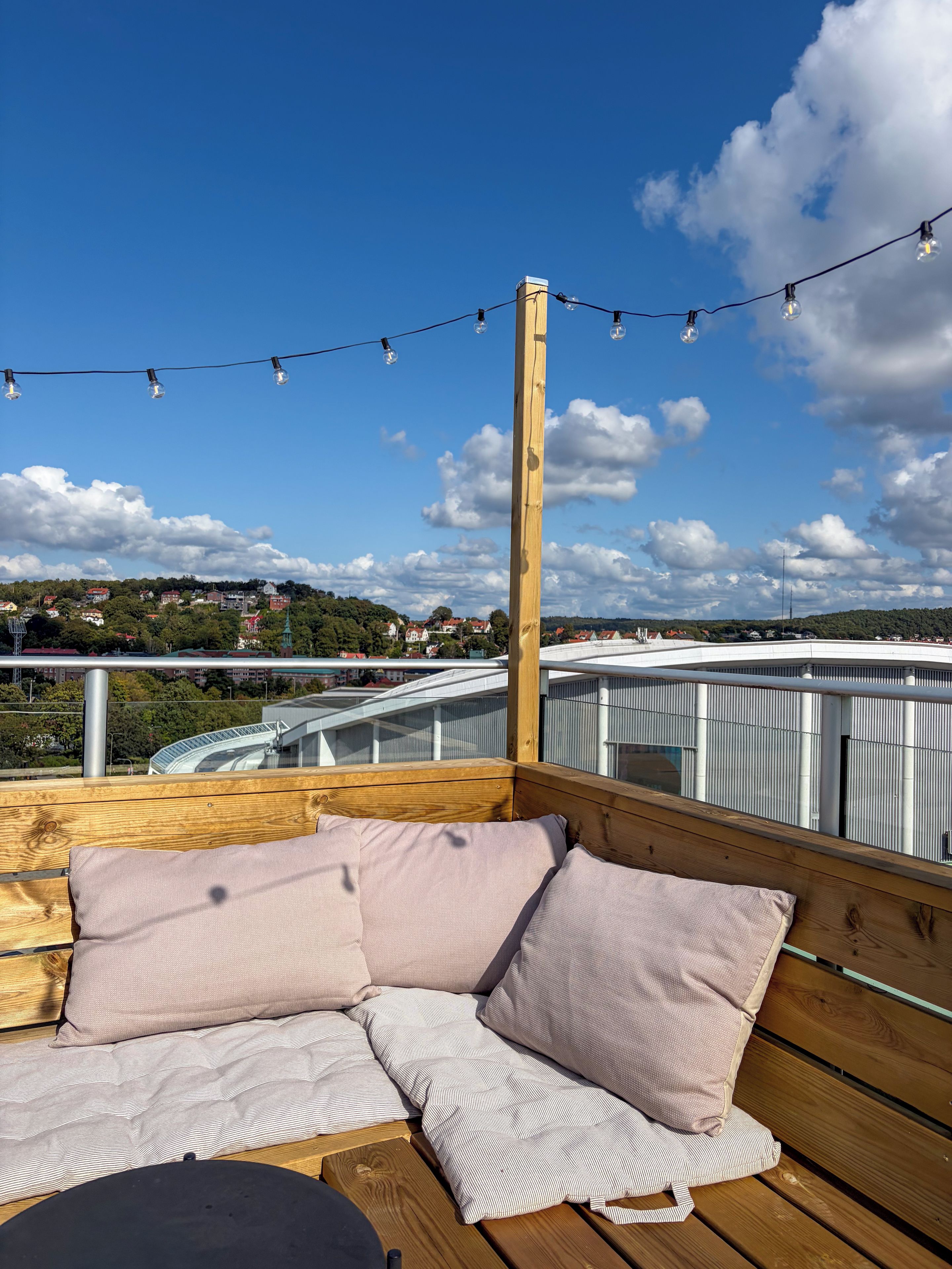 Um deck de madeira com um banco e almofadas, com vista para um horizonte urbano com edifícios e uma ponte.