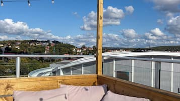 Una terraza de madera con un banco y cojines, con vistas a un paisaje urbano que incluye edificios y un puente.