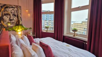 A hotel room with a large bed, red curtains, a view of buildings, and a painting on the wall.