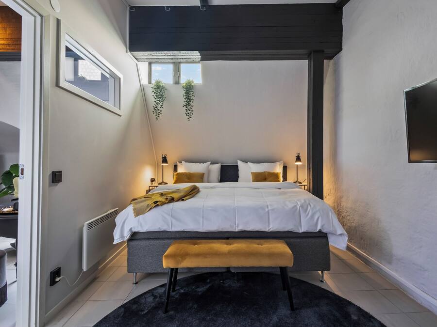 Un dormitorio moderno con cama, mesitas de noche, un banco, un radiador y una ventana.