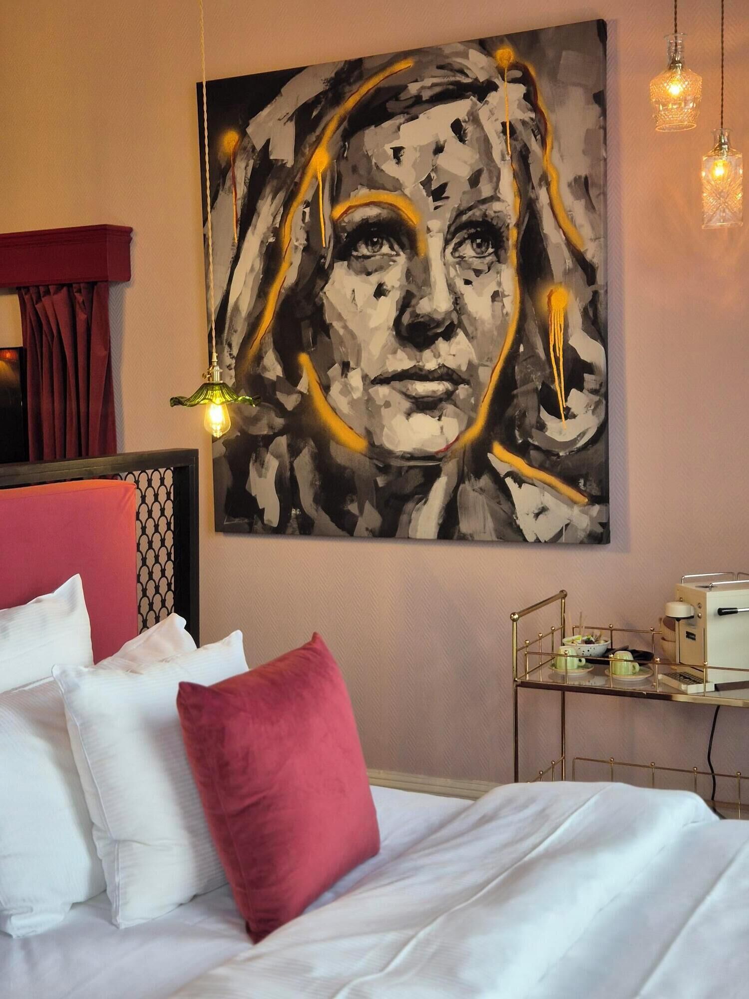 A bedroom with a bed, pillows, a bedside table with a lamp, and a wall-mounted painting.