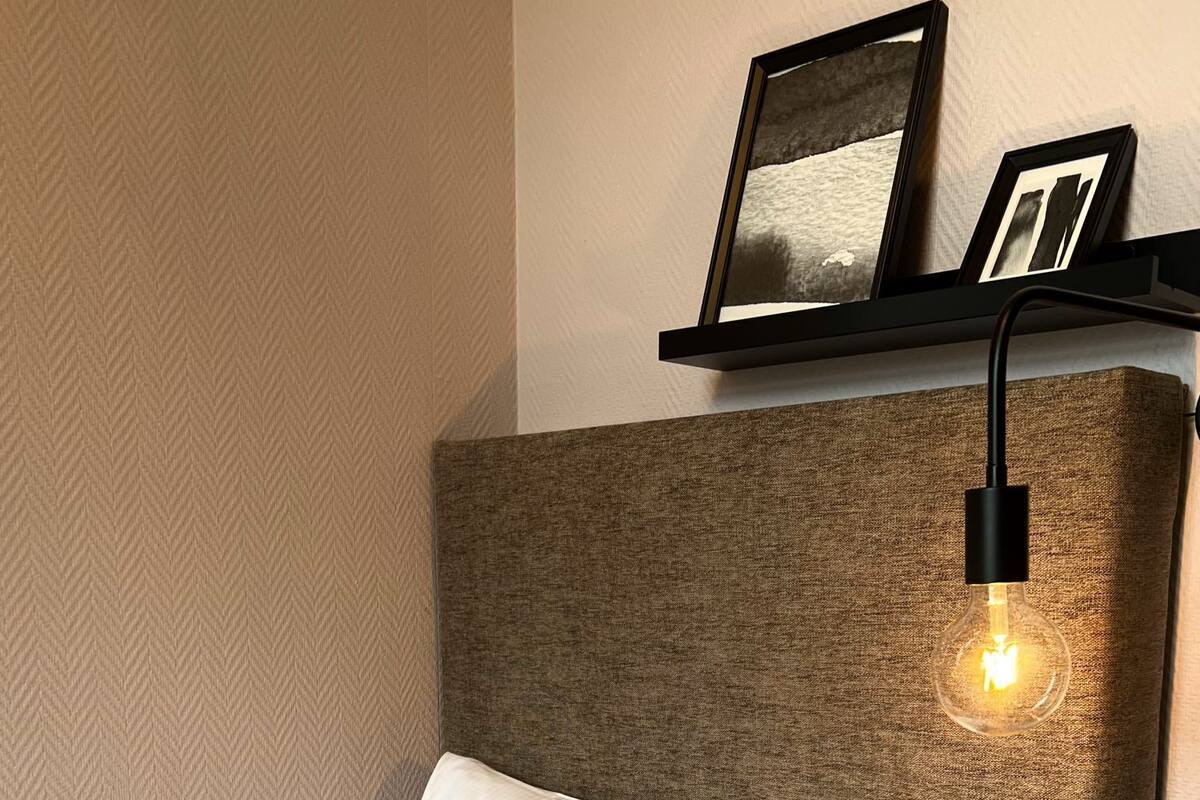 Uma cama bem arrumada com almofadas brancas e verdes estampadas, cabeceira com tecido texturizado e luminária fixada na parede.