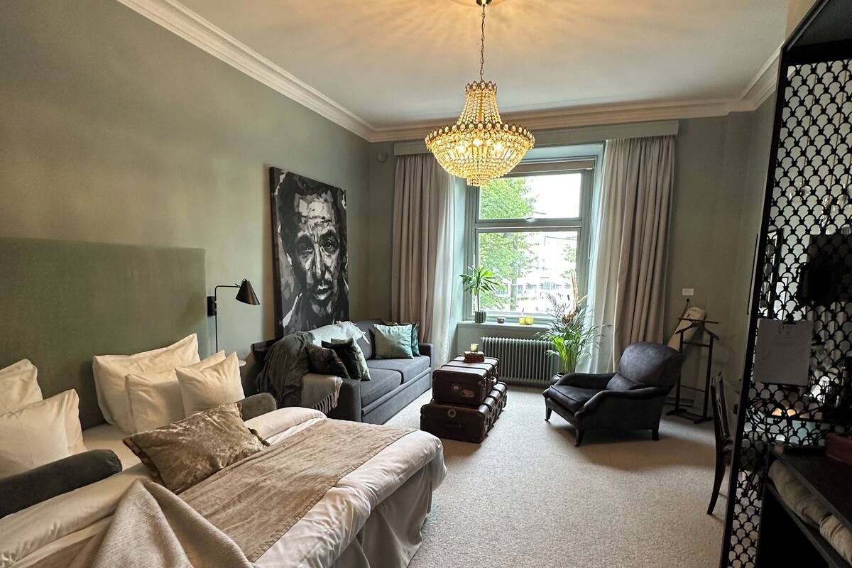 Um quarto espaçoso com uma cama grande, um sofá, uma poltrona e uma escrivaninha. Há uma janela com cortinas e um candelabro pendurado no teto.