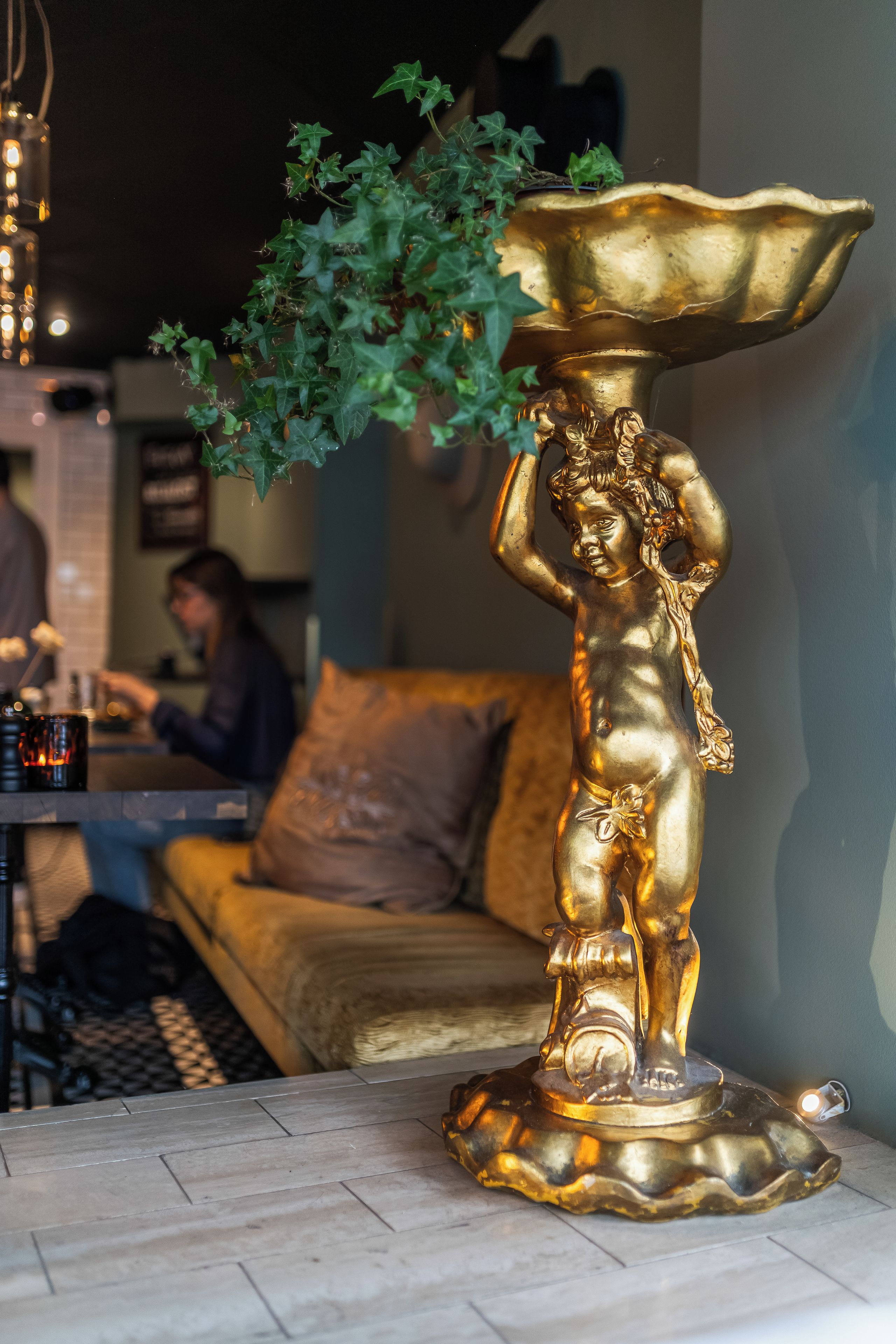 Una urna decorativa dorada con una planta encima, ubicada sobre un suelo de baldosas en una habitación con un sofá y una mesa con bebidas.