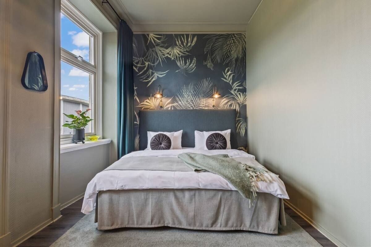 Quarto com cama, janela com cortinas, parede com padrão de folha e mesa de cabeceira com planta.