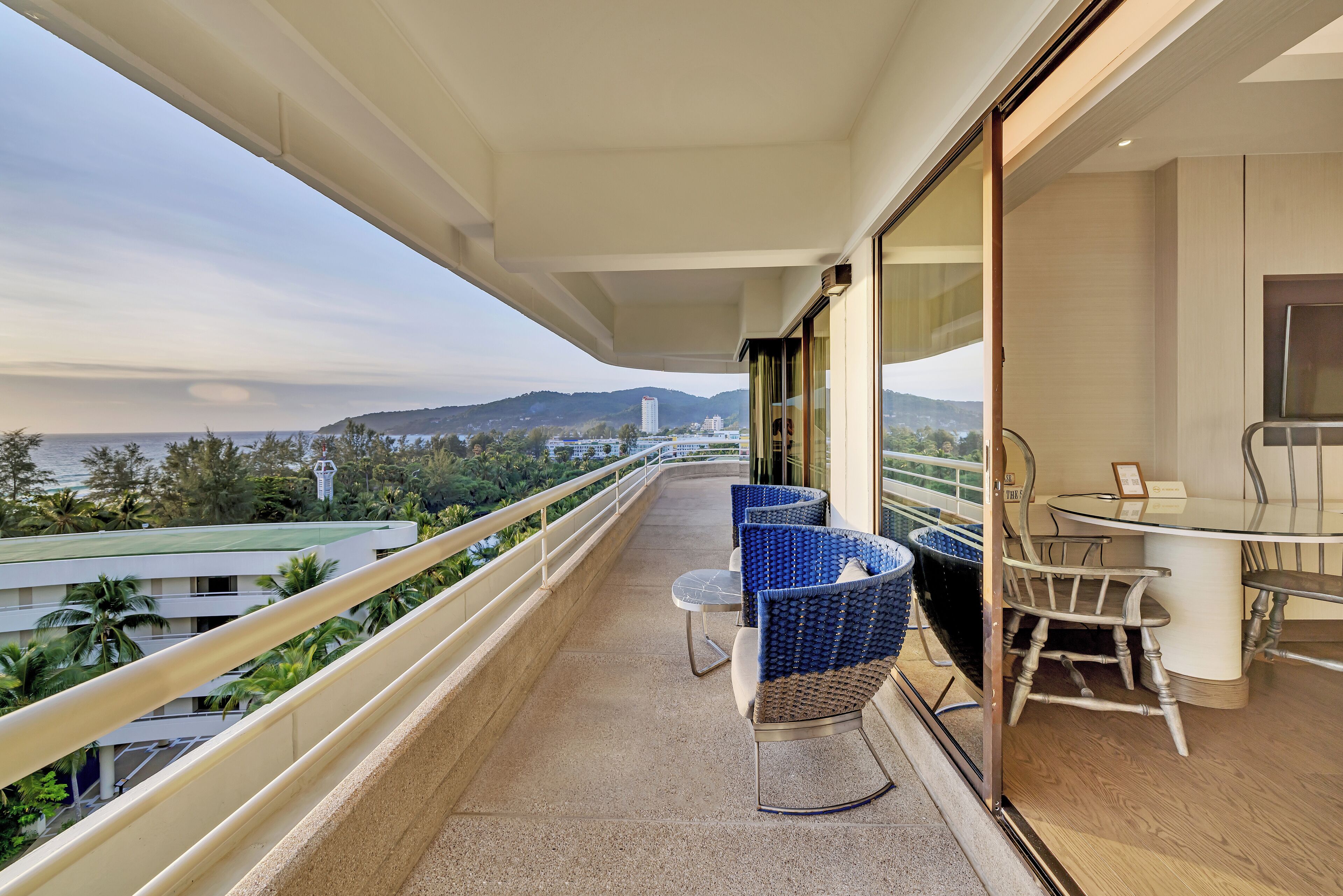 A balcony with a view of the sea and hills, furnished with blue and white chairs, and a table.