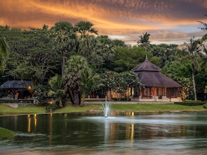 A traditional-style building with a thatched roof, surrounded by lush greenery and a calm pond with a fountain. - Pullman Phuket Karon Beach Resort (Karon)