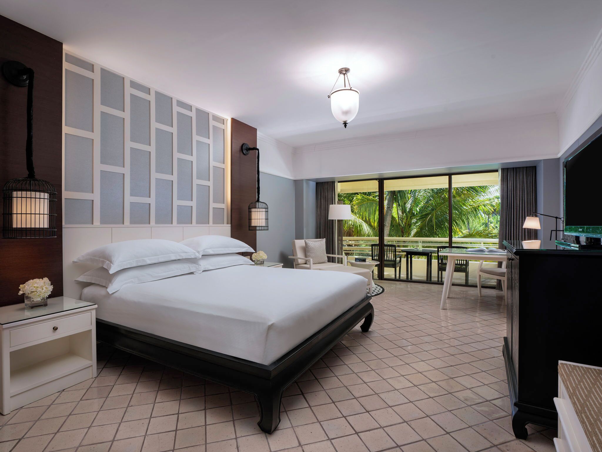 Une chambre d’hôtel comprenant un lit, des tables de chevet, un bureau, une chaise, une télévision et un balcon avec vue sur les palmiers.
